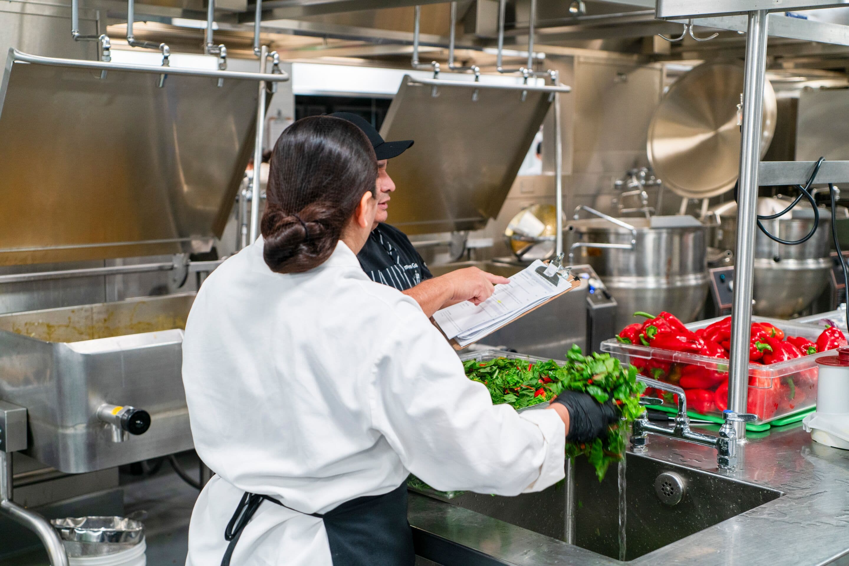 Chef manager gives clear directions to a colleague who washes fresh vegetables. Quality assurance fosters healthy, safe kitchen. 
