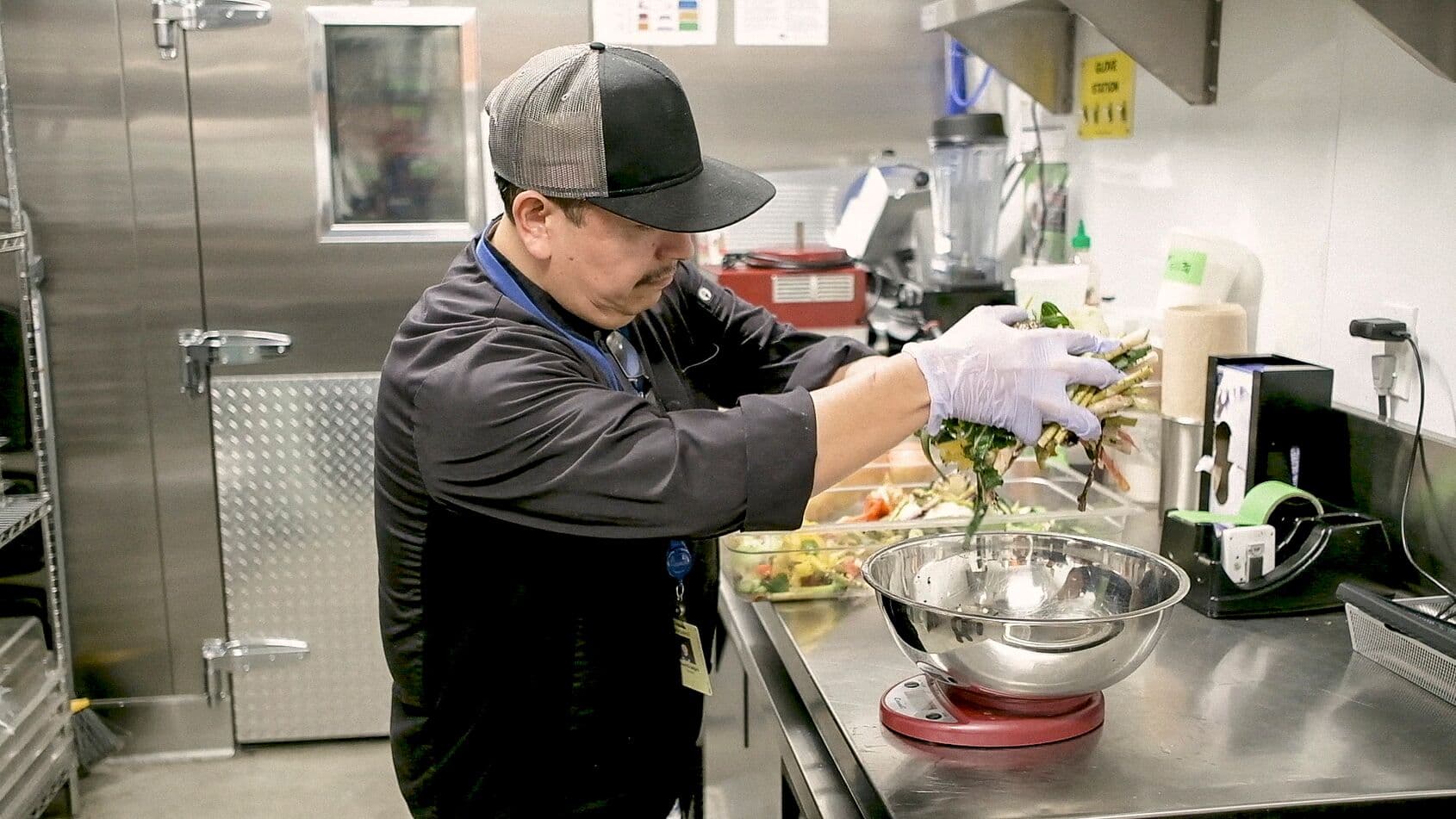 Chef weighs vegetable trimmings on a scale in a workplace dining kitchen to reduce food waste and practice sustainability.